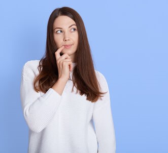 Young pensive girl with dark hair, keeps fore finger near corner of lips, looking aside while pondering on idea, has doubts, wears casual white shirt, models over blue background.